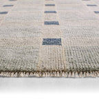 Cera Tiles hand-knotted wool rug in muted grey and cream with blue geometric accents and fringe.