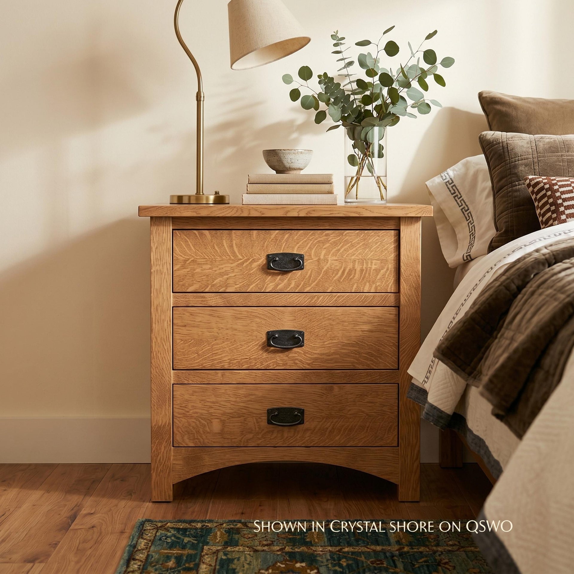 Claremont Wide Mission Nightstand in light oak wood with three drawers next to a bed.