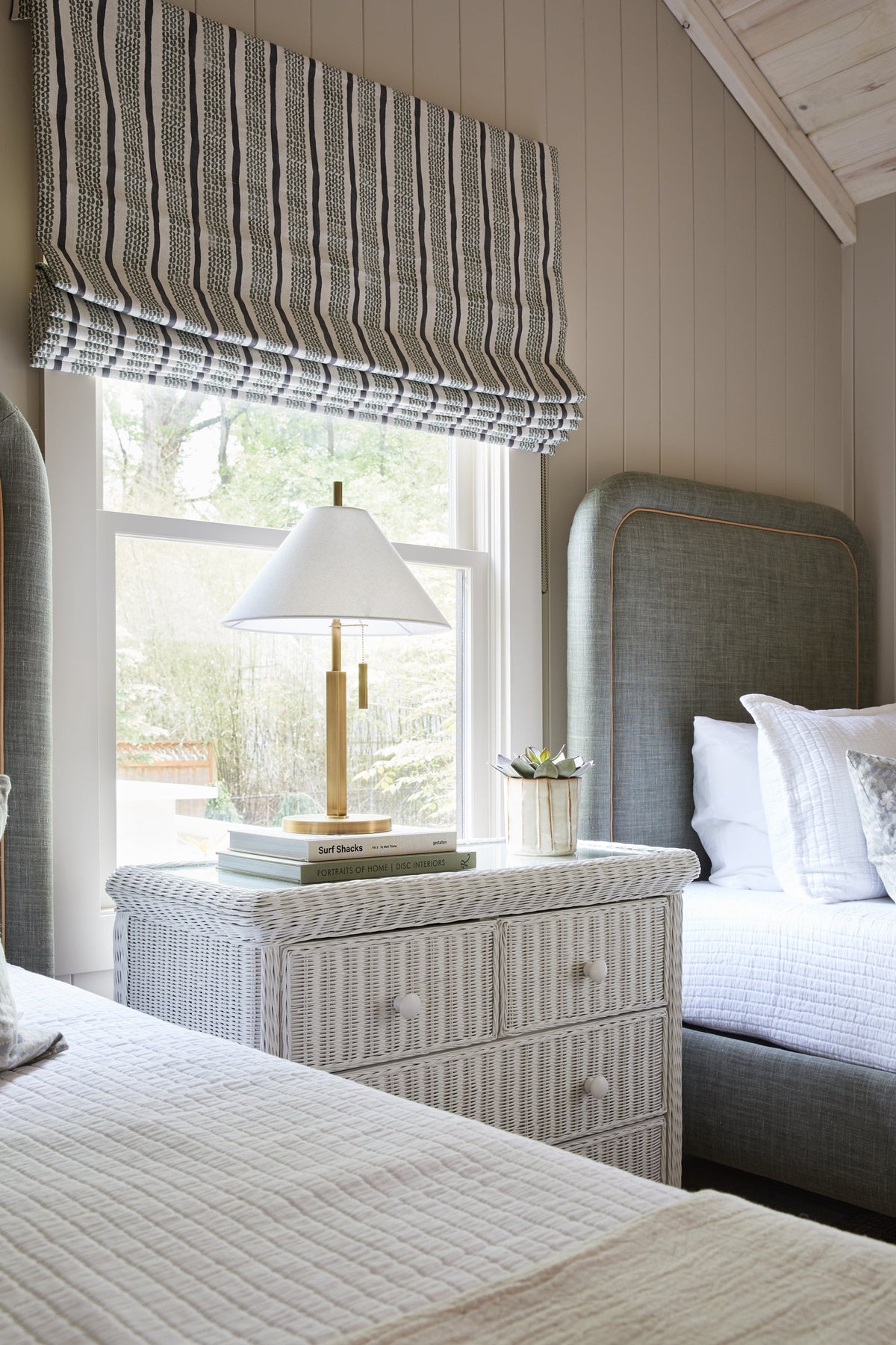 Clic brass table lamp with off-white linen shade on a white wicker nightstand between two beds.