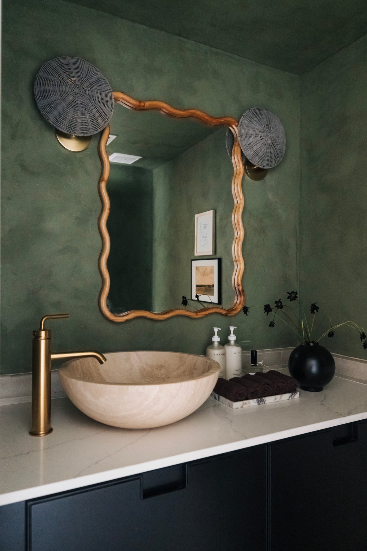 Destiny gray wicker and aged brass wall sconces flanking a wavy wood mirror in a green bathroom.