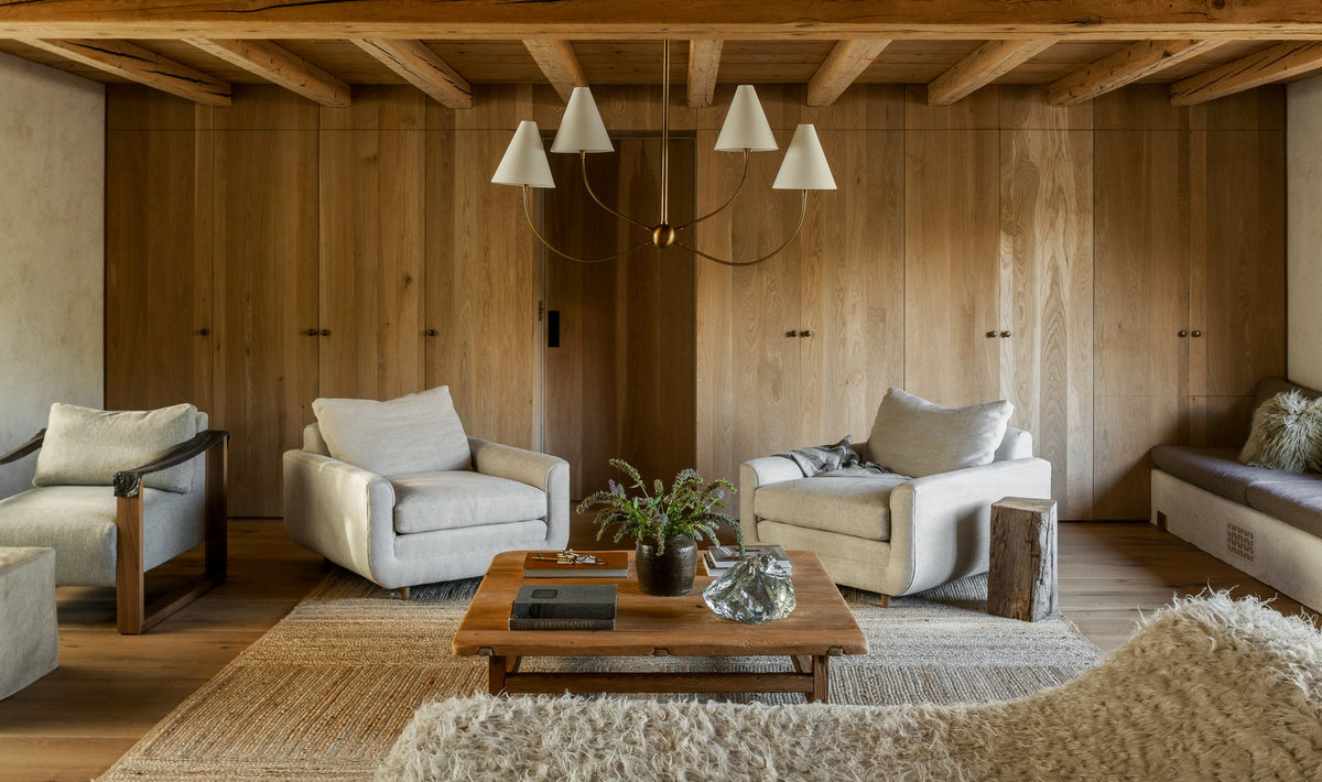 Evermore linear brass chandelier with off-white linen shades in a rustic wood-paneled living room.