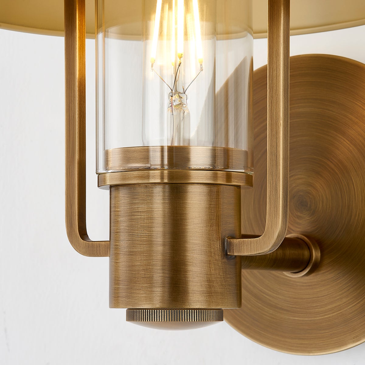 Patina brass Kindle wall sconce with clear glass housing and glowing filament bulb.