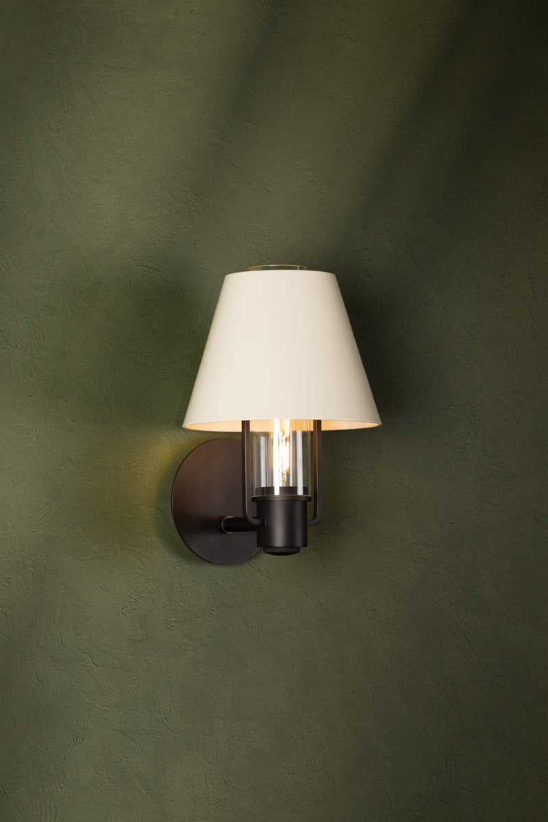 Kindle wall sconce with bronze metalwork, clear glass, and a tapered soft sand shade on a green wall.