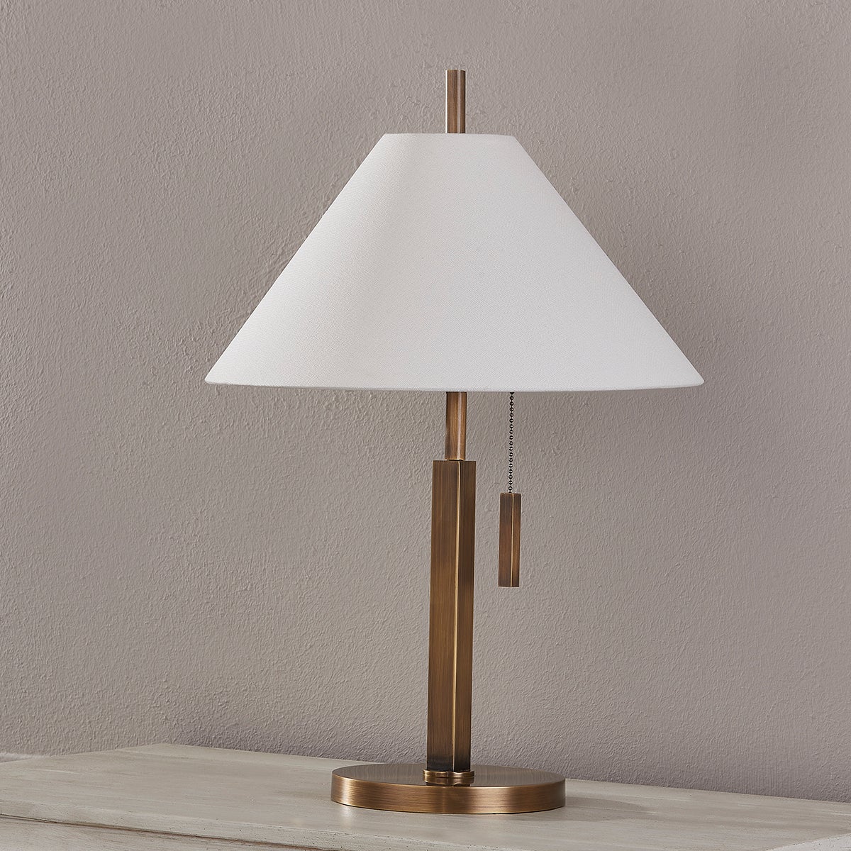Clic table lamp with a patina brass base, off-white linen shade, and a pull chain switch.