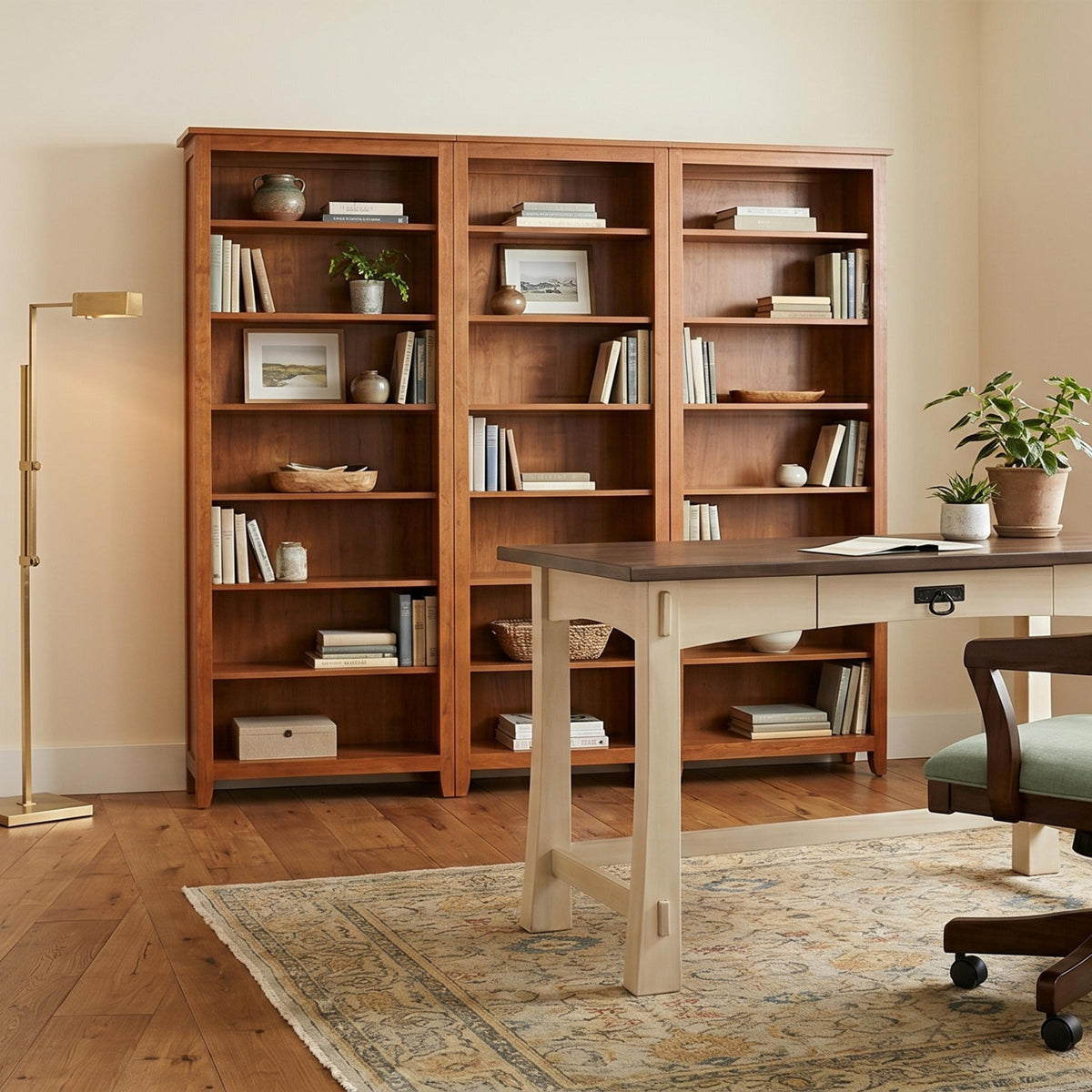 Three-section cherry wood Shaker bookcase filled with books and decor behind a home office desk.