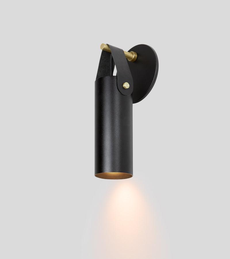 Spero minimalist black cylinder wall sconce with leather strap and brass accents.