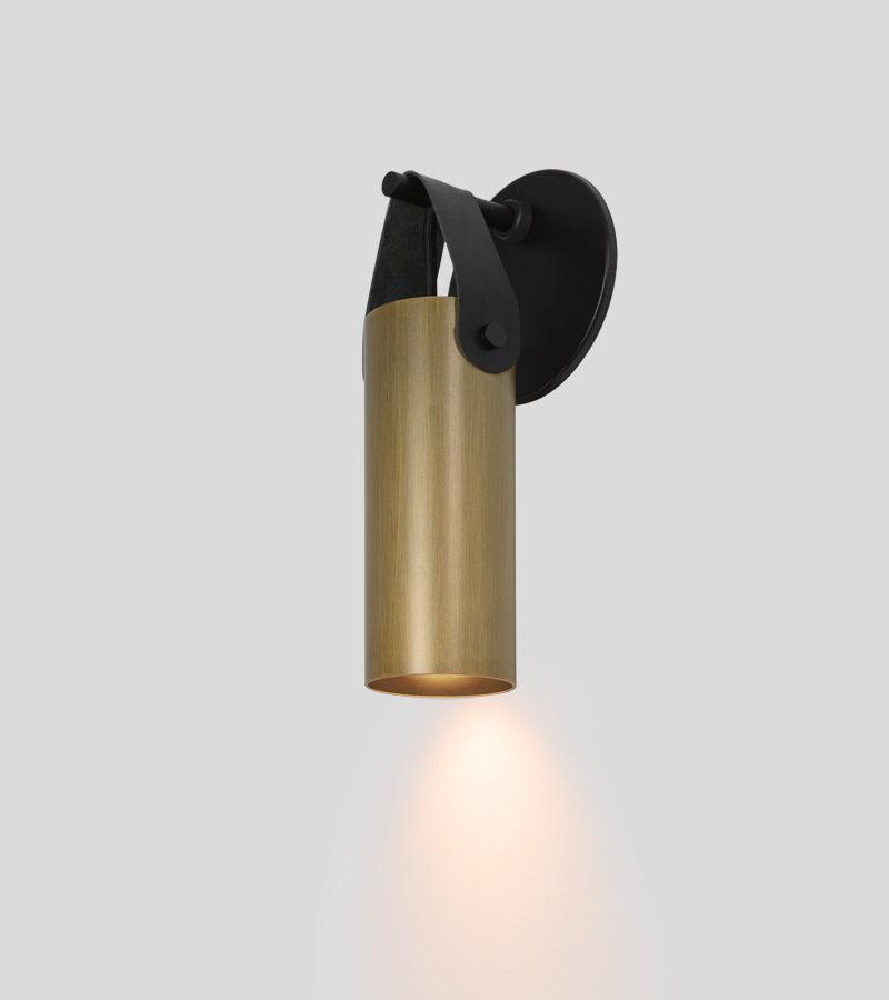 Spero sconce in brushed brass and black leather, casting a warm downward glow.