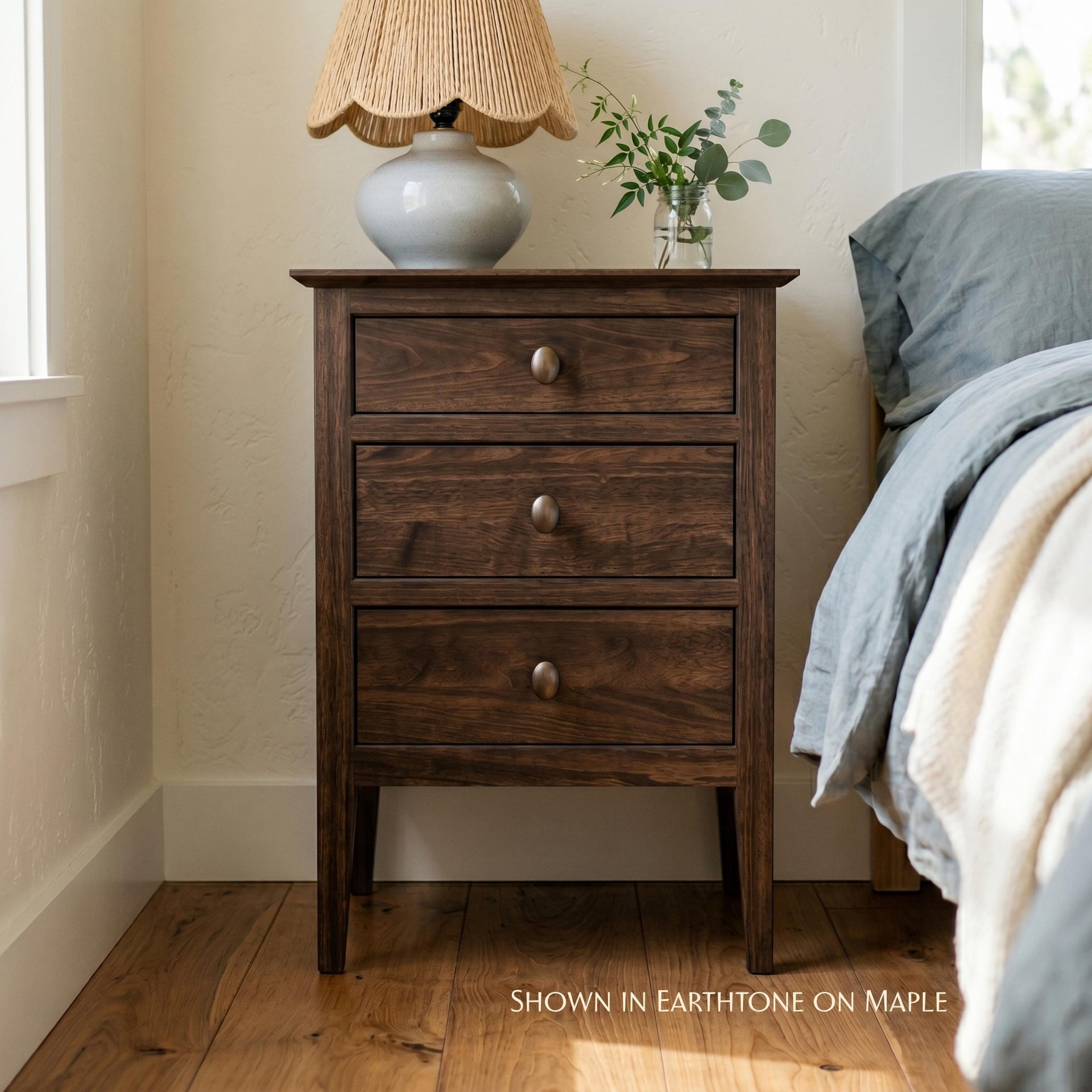 Waterford 3-drawer nightstand in earthtone on maple with rustic brass knobs next to a bed.