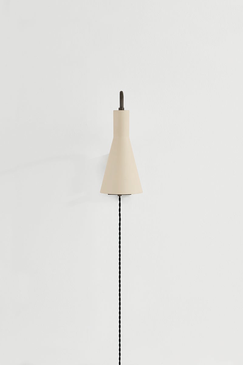 Yvette plug-in sconce in Soft Bisque with a cone shade and black cord on a white wall.