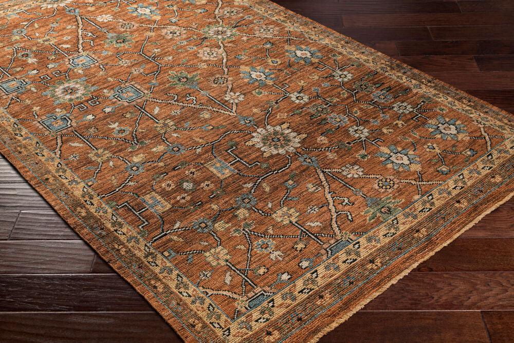 Reign hand-knotted wool rug in rust orange with blue and beige floral patterns on dark wood floor.